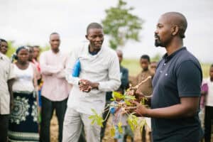 a man holding plants and a group of people around him watch closely while he speaks