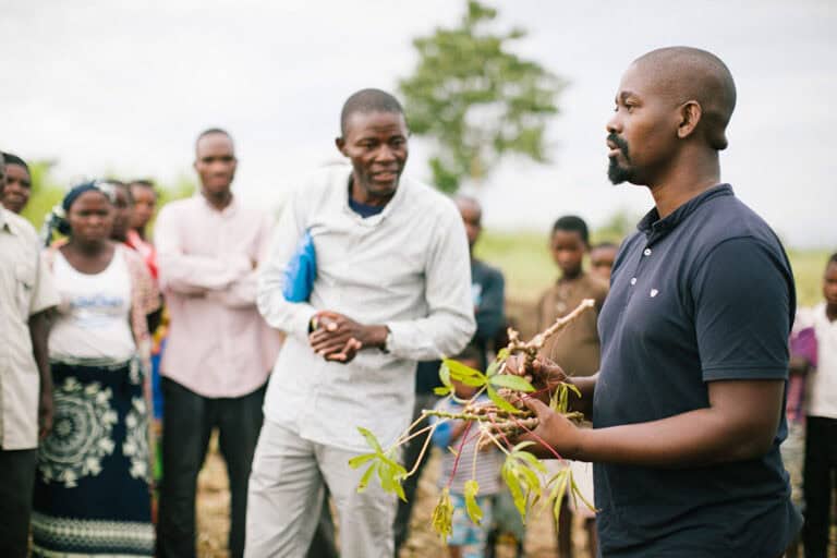 a man holding plants and a group of people around him watch closely while he speaks