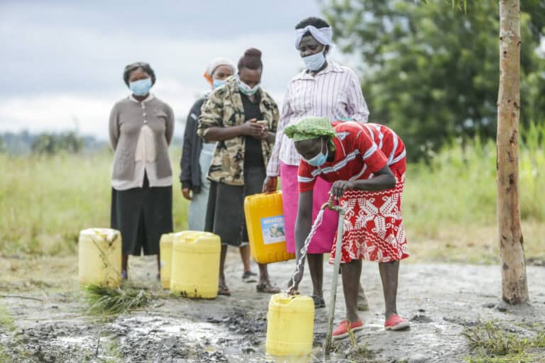 People wearing face masks collect water from a community tap while observing physical distancing outdoors.