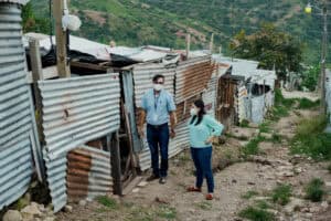 Two people wearing face masks stand beside corrugated metal homes on a hillside road.