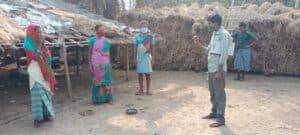 Masked community members in a rural village receive information from an outreach worker holding a clipboard