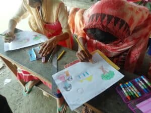 Two Bangladeshi students sitting behind rustic wood desktop, drawing colorful pictures.