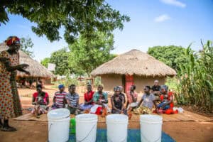 A group of adults seated outdoors near homes while a facilitator speaks beside containers used for a group activity.