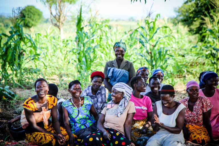 A group of people sitting outdoors together near tall crops, wearing colorful clothing and head coverings.