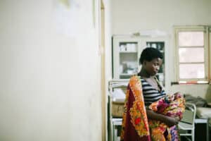 Adult caregiver holding an infant inside a small medical clinic room with shelves and a hospital bed in the background.