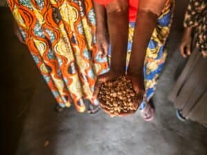 Hands holding a small pile of dried beans, with several people standing close together in the background.