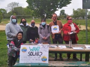 Group of community members standing behind a table with solar information signs at an outdoor park event.