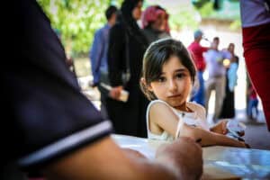 Child sitting at a table while an adult writes on paper, with a group of people gathered outdoors in the background