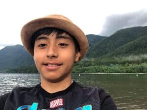Young person wearing a hat near a lake, with forested mountains and cloudy sky in the background.