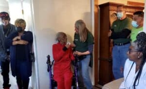 An older adult seated with a walker inside a home, surrounded by standing volunteers wearing masks.