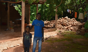Two people stand outside a damaged home with piles of fallen bricks and a simple structure nearby after an earthquake.