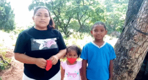 Three people standing outdoors near trees, including an adult and two children, facing the camera.