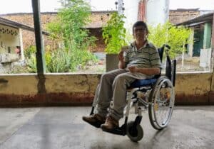 Person using a wheelchair sits on a porch holding a small card, with plants and buildings visible in the background.