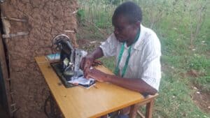 Person sewing fabric on a manual sewing machine outdoors beside an earthen wall.