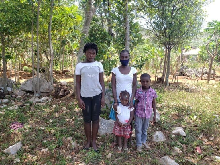 Four people stand outdoors among trees and rocks in a rural area, facing the camera.