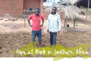 Two people stand in a farmyard near brick buildings and a thatched shelter, with dried crop residue covering the ground.