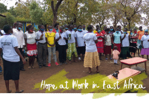 A group of people stand outdoors during a community meeting, with one person speaking at the front with overlaying script, Hope at Work in East Africa.