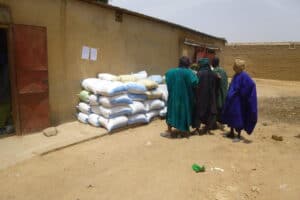 People standing near stacked sacks of food outside a building in a dry, rural setting.