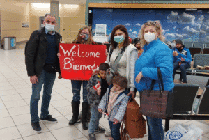 Group of adults and children wearing masks stand in an airport terminal holding a “Welcome” sign, with seating visible behind them.
