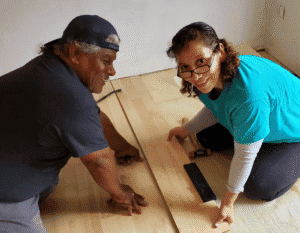 Two people kneel indoors installing wooden flooring using hand tools during home repairs.