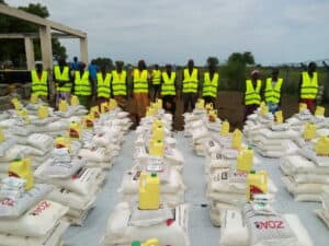 Group of community members standing behind stacks of food aid supplies, including bags of grain, beans, and cooking oil.