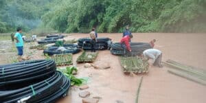 People working in shallow river water, assembling coiled pipes and bamboo materials for a village water system.