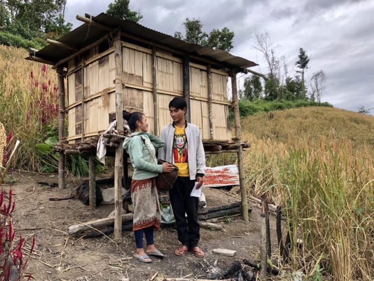 Two people standing beside a small raised wooden structure in a hillside farming area with tall grasses and fields nearby.