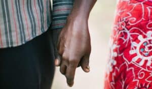 Close-up of two people holding hands, showing their arms and clothing against a blurred outdoor background.