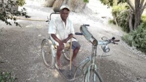 Man sitting in a wheelchair with a bicycle in a rural Mozambique setting.