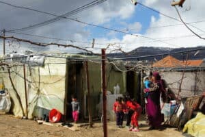 Several adults and children stand outside a makeshift shelter with plastic sheeting and containers in a refugee settlement.