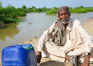 Older adult seated beside a large blue water container near floodwater, with green vegetation in the background.