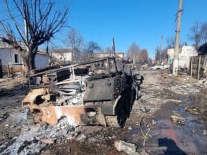 Burned-out armoured vehicle in the middle of a damaged residential street, with destroyed buildings, debris, and utility poles nearby.