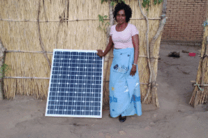 Person standing beside a solar panel outside a woven reed structure in a rural setting.