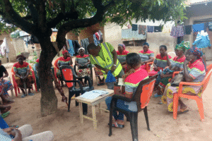 People seated outdoors in a circle while one person records notes at a small table during a community meeting.
