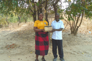 Two adults standing outdoors, holding a small wooden cash box together beneath trees in a rural setting.