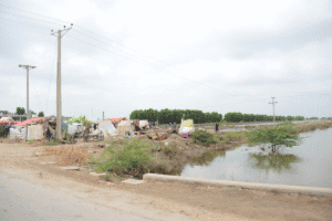 Temporary shelters set beside floodwater along a rural roadside, with tents, people, power lines, and trees in the background.
