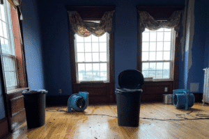 Interior room with water on wooden floors, drying fans, bins, and buckets set up near tall windows.