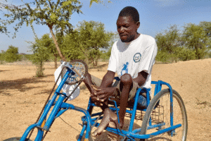 Person seated on a hand‑powered wheelchair outdoors in a dry, sparsely vegetated landscape with scattered trees.