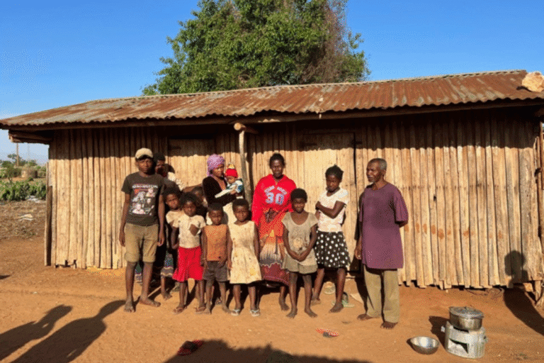 A large family stands outside a simple wooden house with a tin roof in a rural setting.