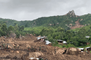 People walking across a muddy landslide area near damaged homes and trees on a hillside after severe flooding.