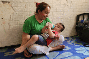 Adult seated on the floor feeding a toddler with a bottle inside a modest room with worn walls.
