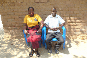 Two adults seated on blue chairs outside a brick building, facing the camera in daylight.