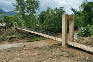 Suspension-style bridge crossing a river between two rural villages, surrounded by trees and hills