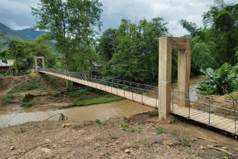Suspension-style bridge crossing a river between two rural villages, surrounded by trees and hills