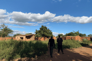 Two people walking along a dirt path past brick houses and greenery under a blue sky in a rural community.