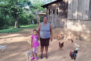 Adult and child standing outside a wooden home in a rural setting with several chickens nearby.