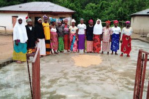 A group of people standing on a fenced concrete surface with rice spread out to dry near village buildings.