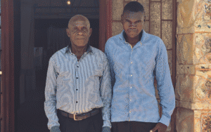 Two men standing outside a building, one elderly and one young, in a community support setting.