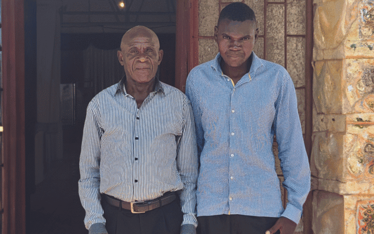 Two men standing outside a building, one elderly and one young, in a community support setting.