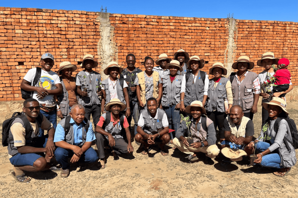 Group of staff standing and kneeling outdoors in front of a brick wall, some holding small plants.